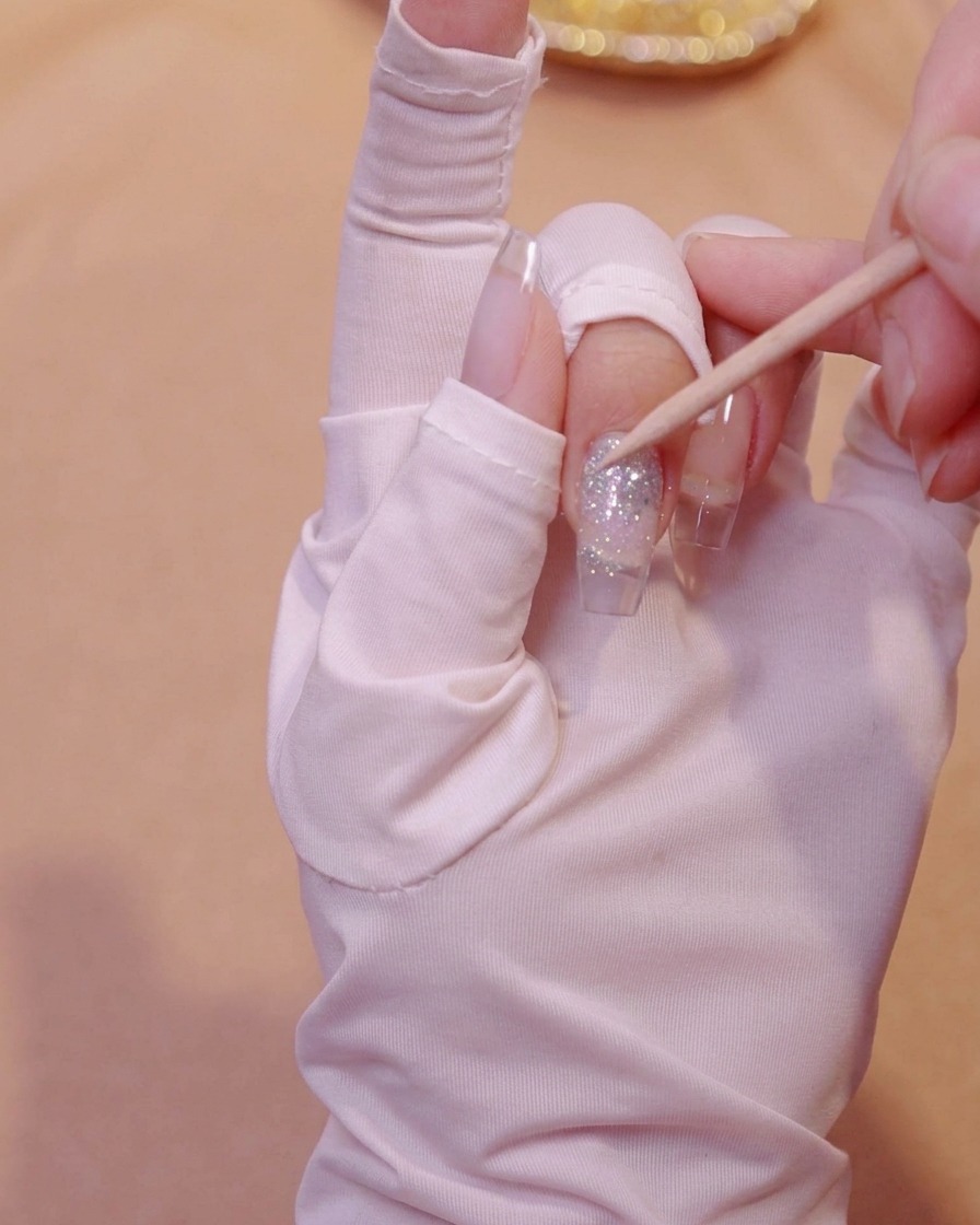 An orangewood stick gently pressing down butterfly sequins embedded in glitter gel on a nail.