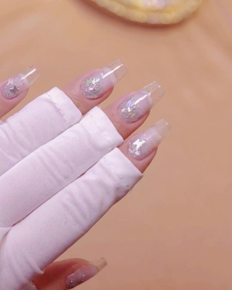 A hand with clear nail extensions and embedded butterfly glitter positioned under a UV lamp.
