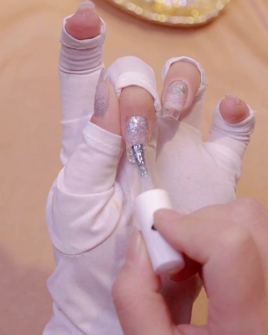 A white-gloved hand using a brush to apply milky white gel polish to the tip of a fingernail.