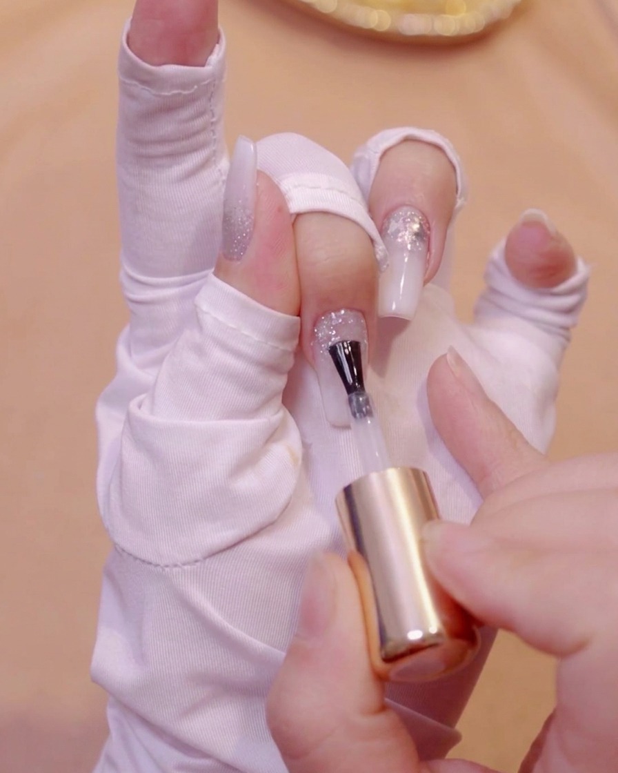 A brush applying clear leveling gel onto a textured glitter nail surface.