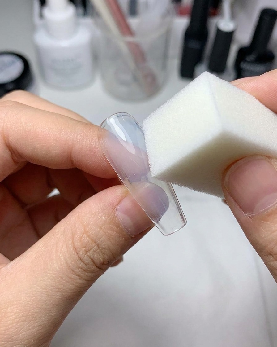 A small makeup sponge pressing light blue gel onto the center of a clear artificial nail.