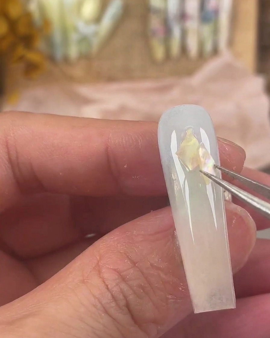 Fine tweezers placing iridescent shell pieces onto the wet gel surface of the nail.