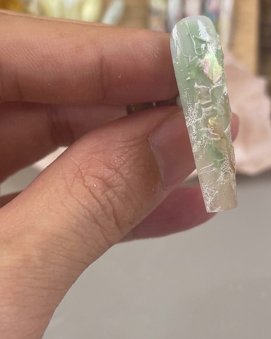 Close-up of a green and clear nail with iridescent shell flakes and white lace patterns stamped across its surface.