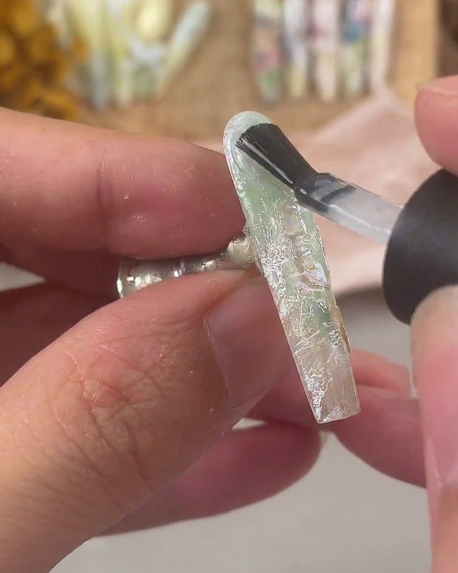 A nail brush applying a thick layer of clear builder gel over a decorated nail with lace and shell flakes.