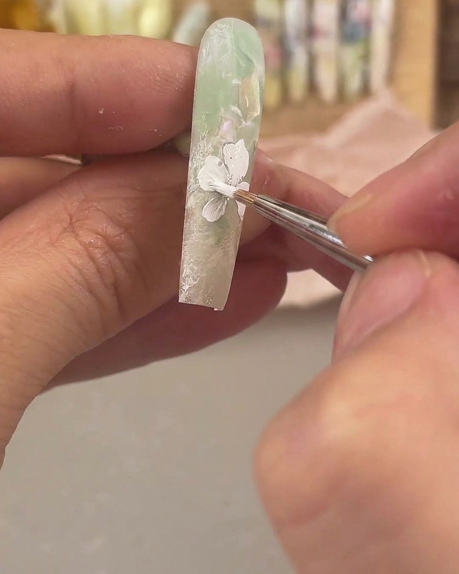 A fine detail brush applying white painting gel to draw a large flower on a matte green and clear nail.