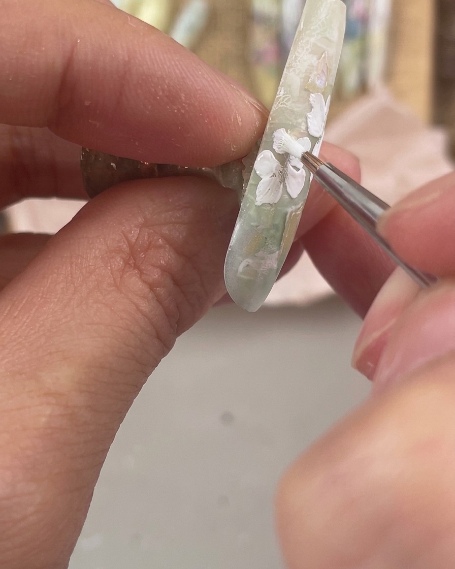 A fine detail brush painting three small white floral petals along the lower side edge of a decorated nail.