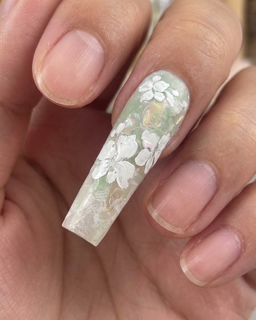 Close-up of a long, sculpted nail showing completed white gel flowers and tiny leaves over a green and shell-flake background.