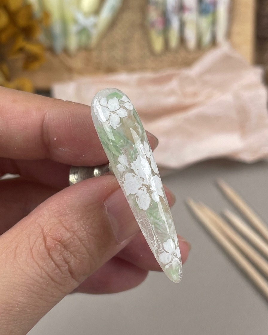 A long, matte-finished nail featuring a white floral design, held on a magnetic nail stand.