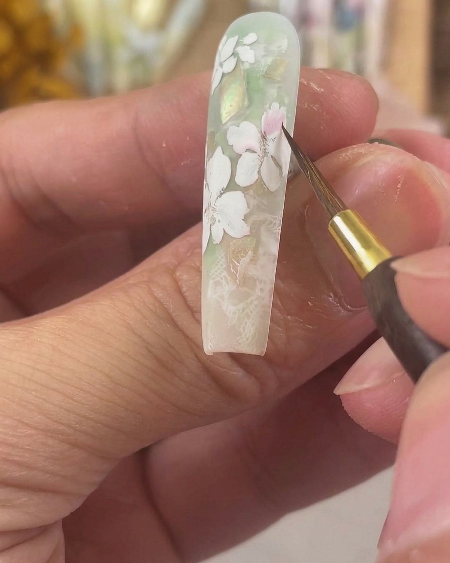 A fine detail brush applying sheer pink watercolor pigment to matte white gel petals on a long sculpted nail.