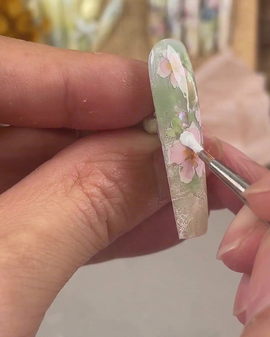 A fine liner brush applying white painting gel to form an extra layer of petals on a pink floral nail art design.