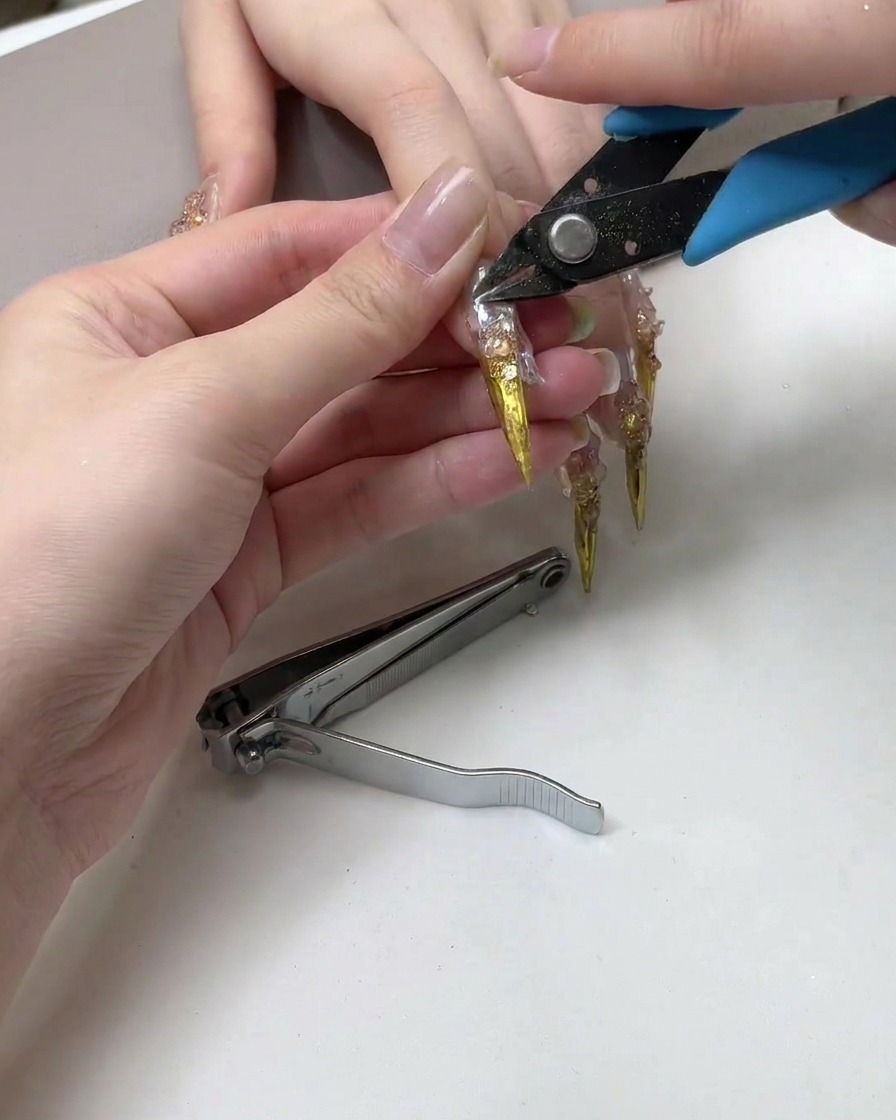 A pair of nail clippers cutting the sharp pointed tip of a long metallic nail extension.