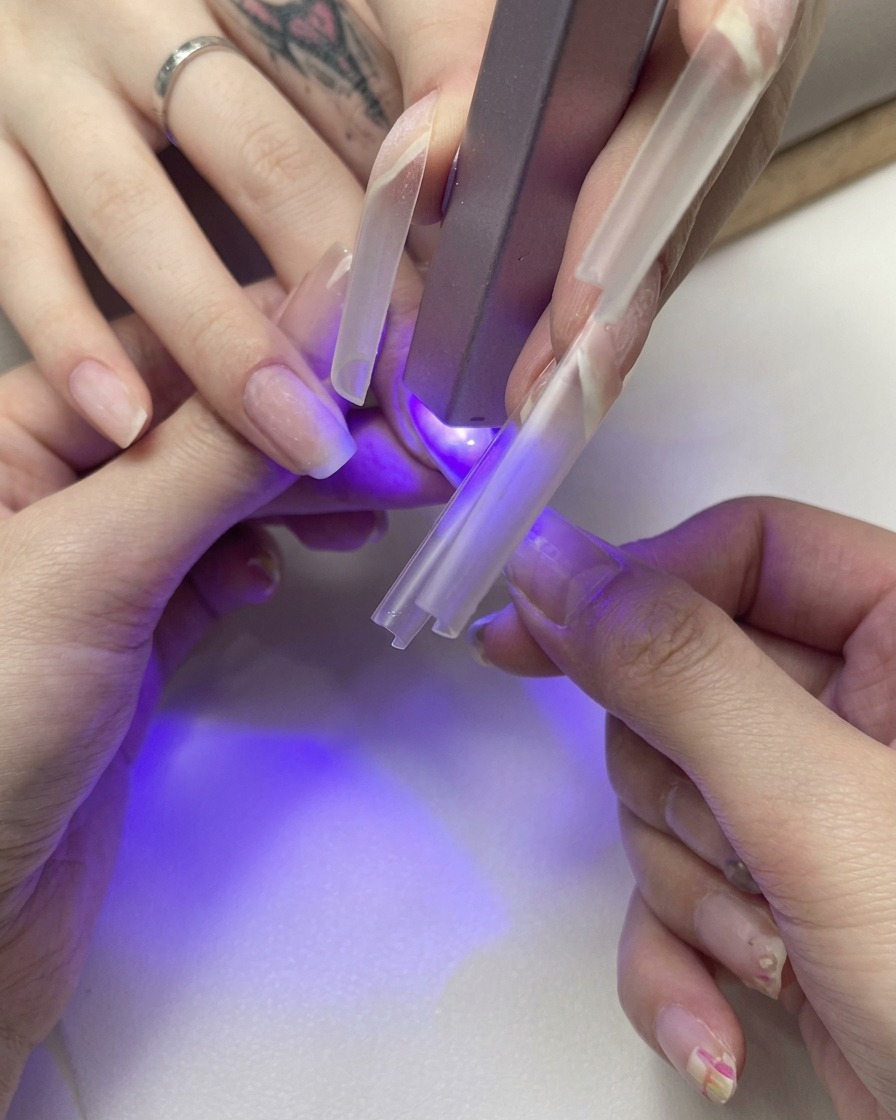 A close-up of a hand holding a clear nail extension tip onto a natural nail while curing it under a small purple UV light.