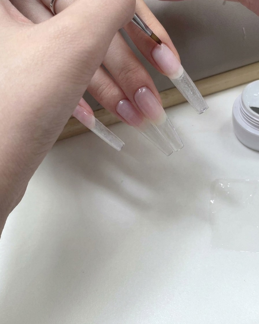A fine nail art brush applying clear gel to the underside of a long transparent nail tip.