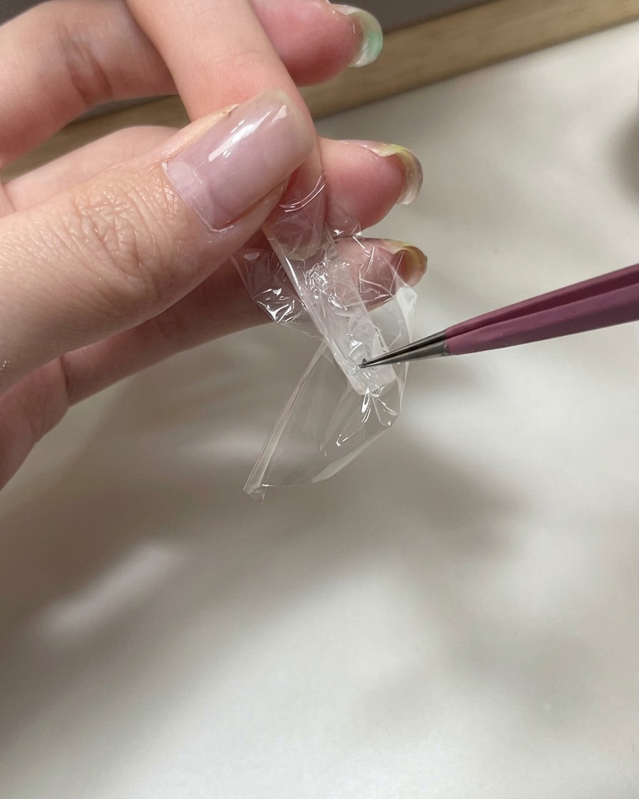 A close-up of tweezers pressing a piece of clear plastic wrap into the underside of a clear nail extension to create a wrinkled texture.
