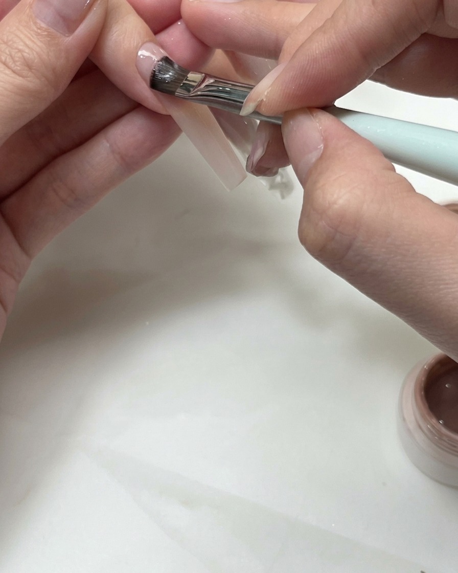 A flat brush applying nude gel to shape a French smile line on a fingernail.
