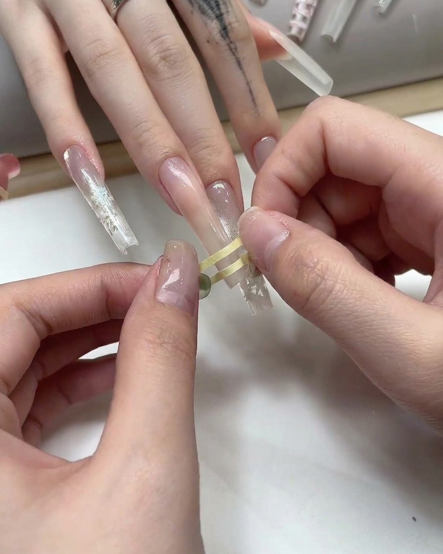Fingers placing thin gold striping tape in a grid pattern onto a nude gel nail.