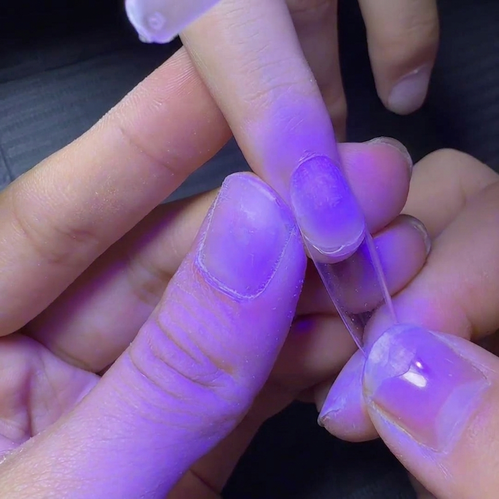 A fingernail with a clear tip being held under a purple UV lamp for a quick flash cure.