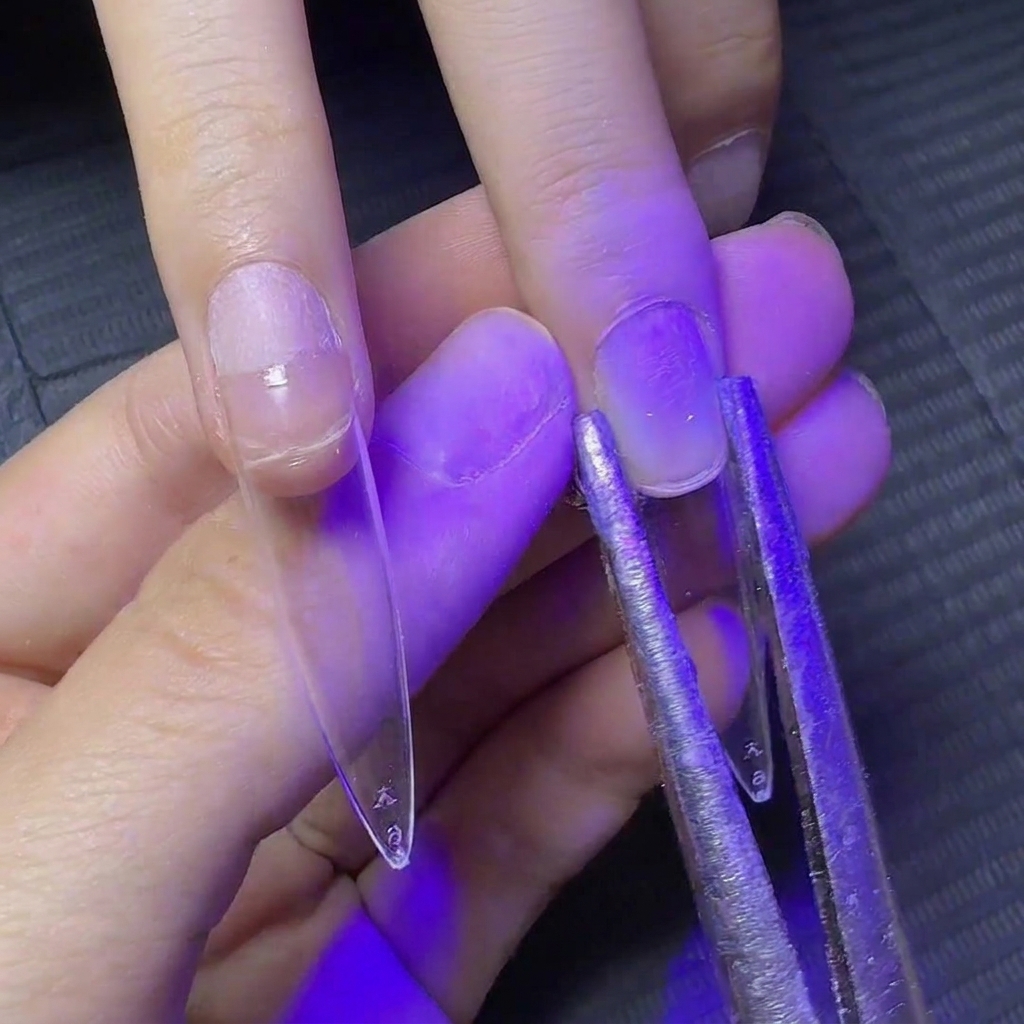 Metal tweezers clamping the sides of a clear extension nail under UV light to shape the curve.