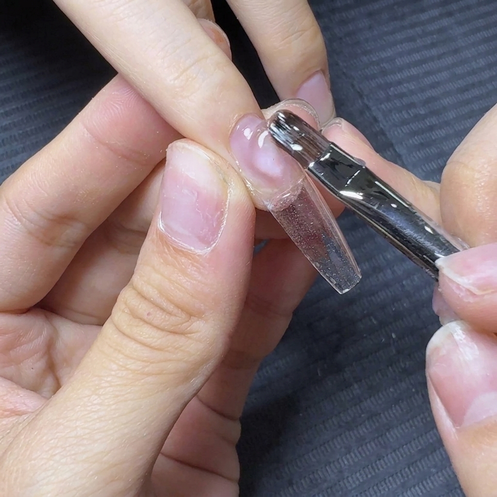 A nail brush floating and dragging clear builder gel down the length of a clear nail extension tip.