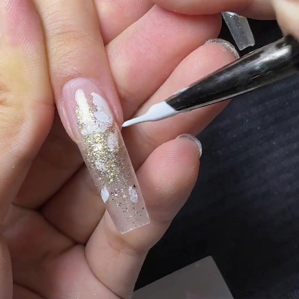A fine liner nail brush painting small white flower petals over a clear nail with gold glitter.