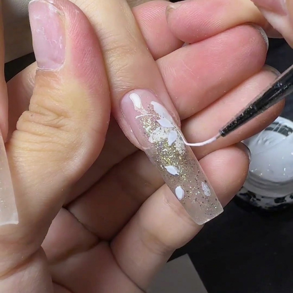 A thin tool pulling stringy white spider gel across a gel nail decorated with white flowers and gold glitter.