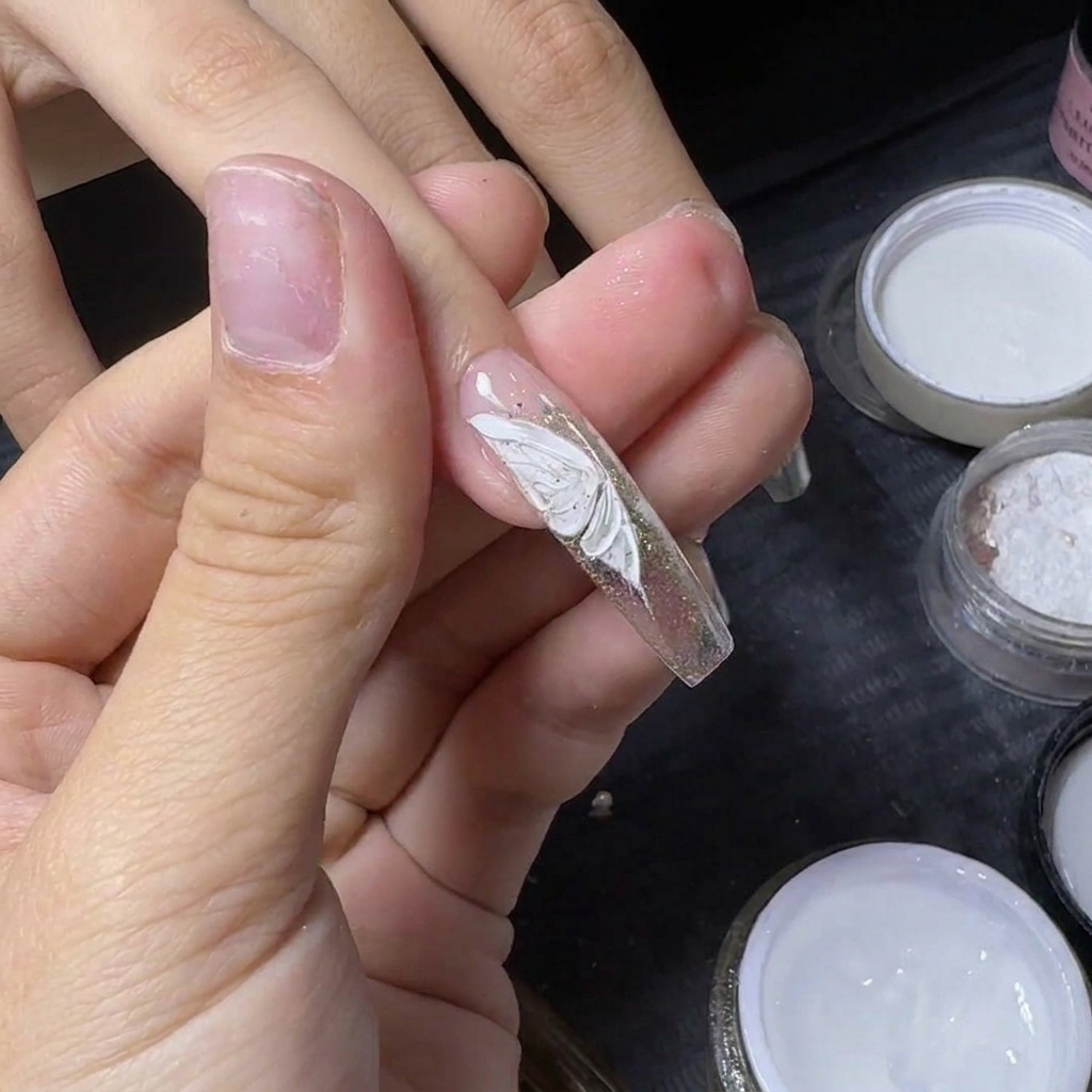 A manicurists thumb holding a nail to attach a small silver rhinestone onto a painted white butterfly.