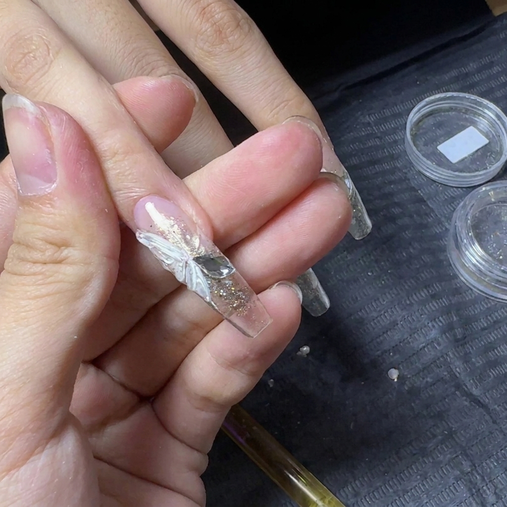 A close-up of a clear nail extension featuring a white 3D butterfly wing with two flat-back rhinestones placed alongside it to create the butterfly bodycite: 1.