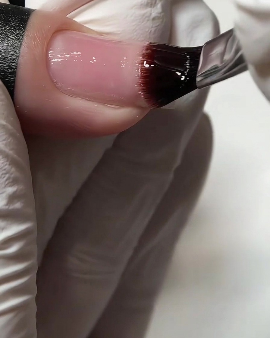 A close-up of a flat gel brush applying clear base coat onto a natural fingernail.