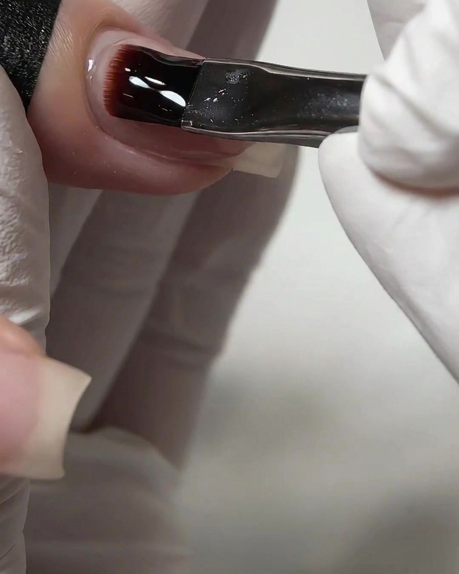 A nail brush applying a thick layer of clear self-leveling builder gel onto a fingernail.