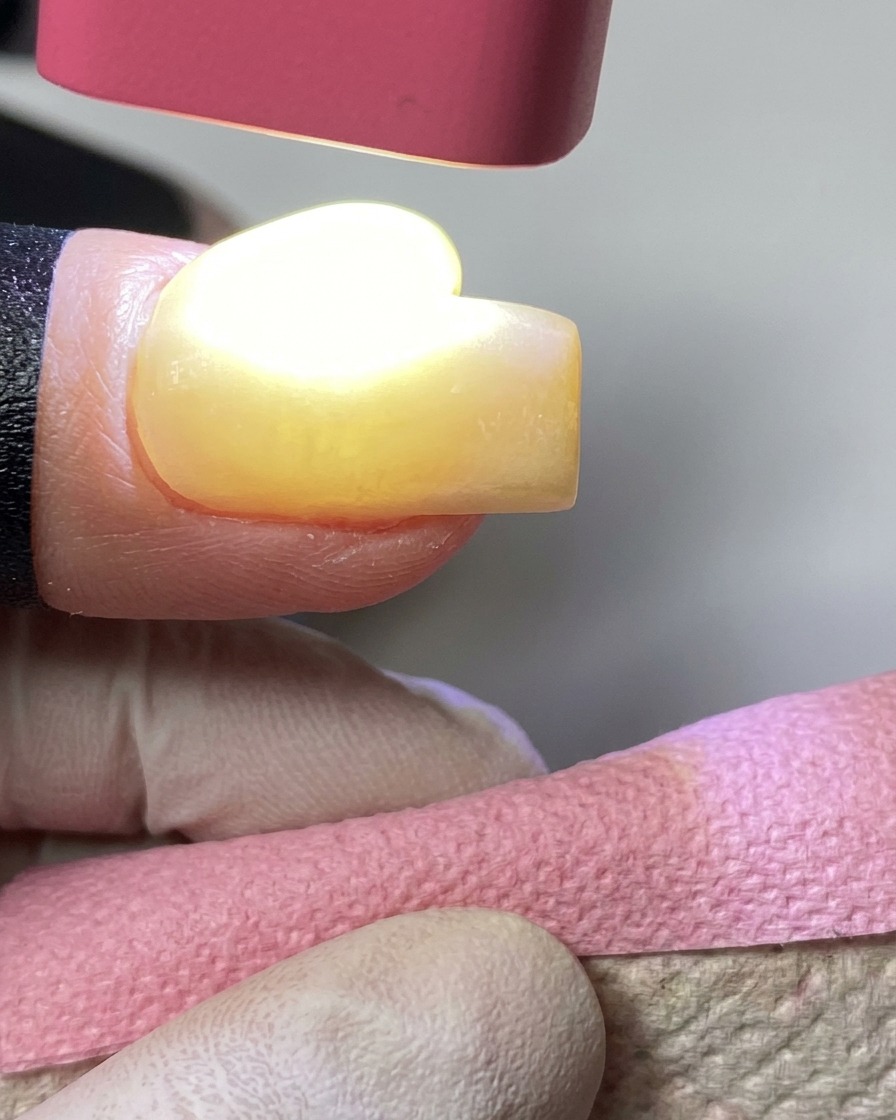 A handheld pink UV lamp curing yellow gel polish on a fingernail.