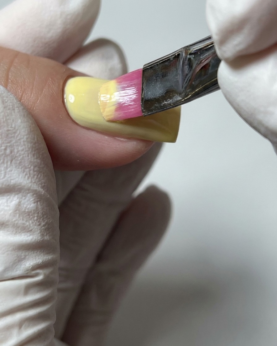 A close-up of a brush applying a second coat of pastel yellow gel polish to ensure complete color coverage.