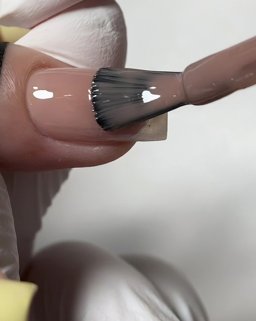 A bottle brush applying a sheer, glossy nude gel polish onto a prepped fingernail.