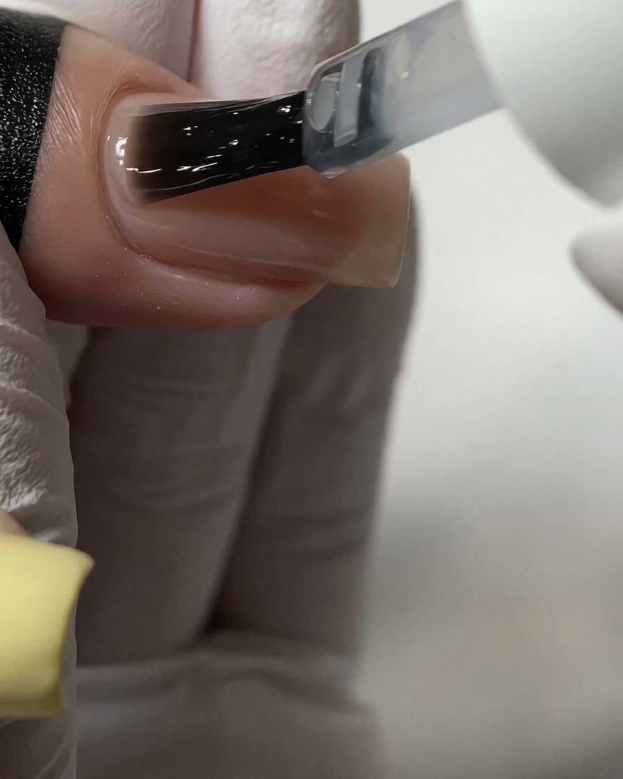 A brush applying a clear top coat over a nude-colored acrylic nail.