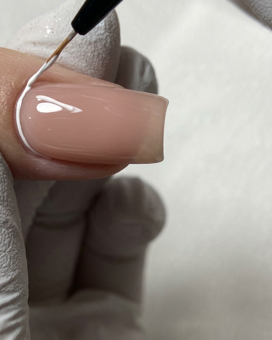 A fine nail art brush drawing white geometric lines near the cuticle on a nude nail.