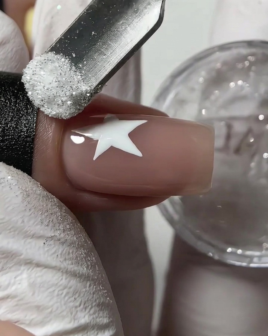 A metal cuticle pusher sprinkling white sugar glitter powder over a wet white star drawn on a nude fingernail.