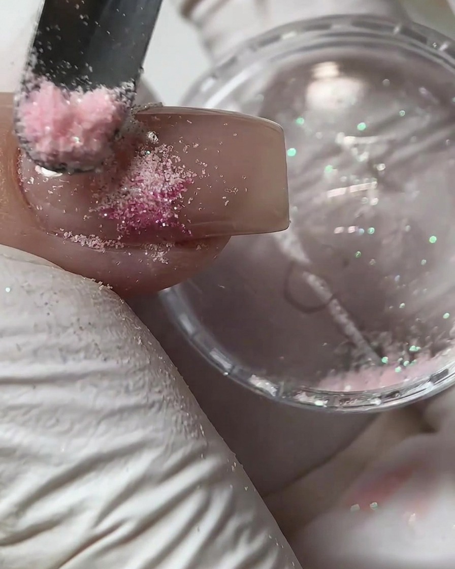A small tool scattering pink sugar powder over a wet gel design on a fingernail.