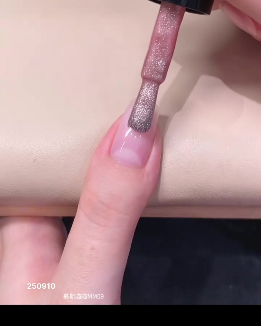 A bottle brush applying a drop of sheer pink magnetic gel polish near the cuticle area of a natural nail.