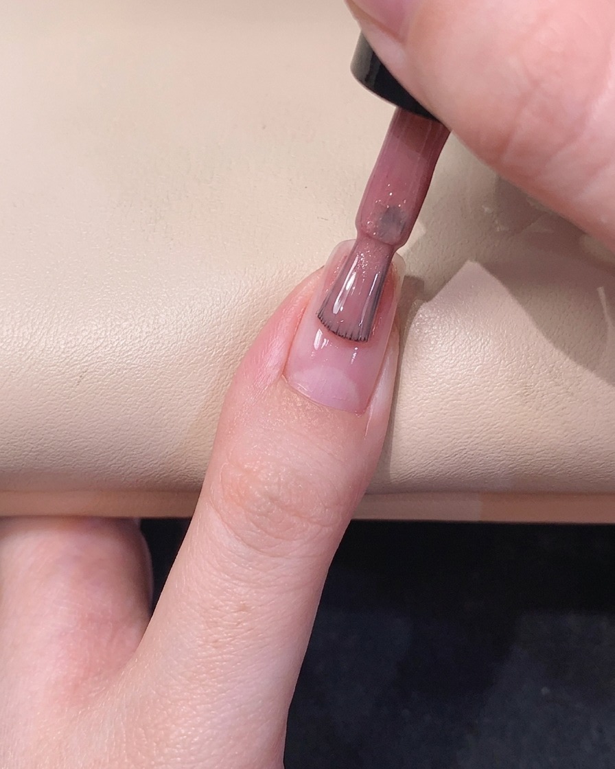 A gel polish brush gliding down the center of the fingernail, distributing a sheer pink sparkly polish.