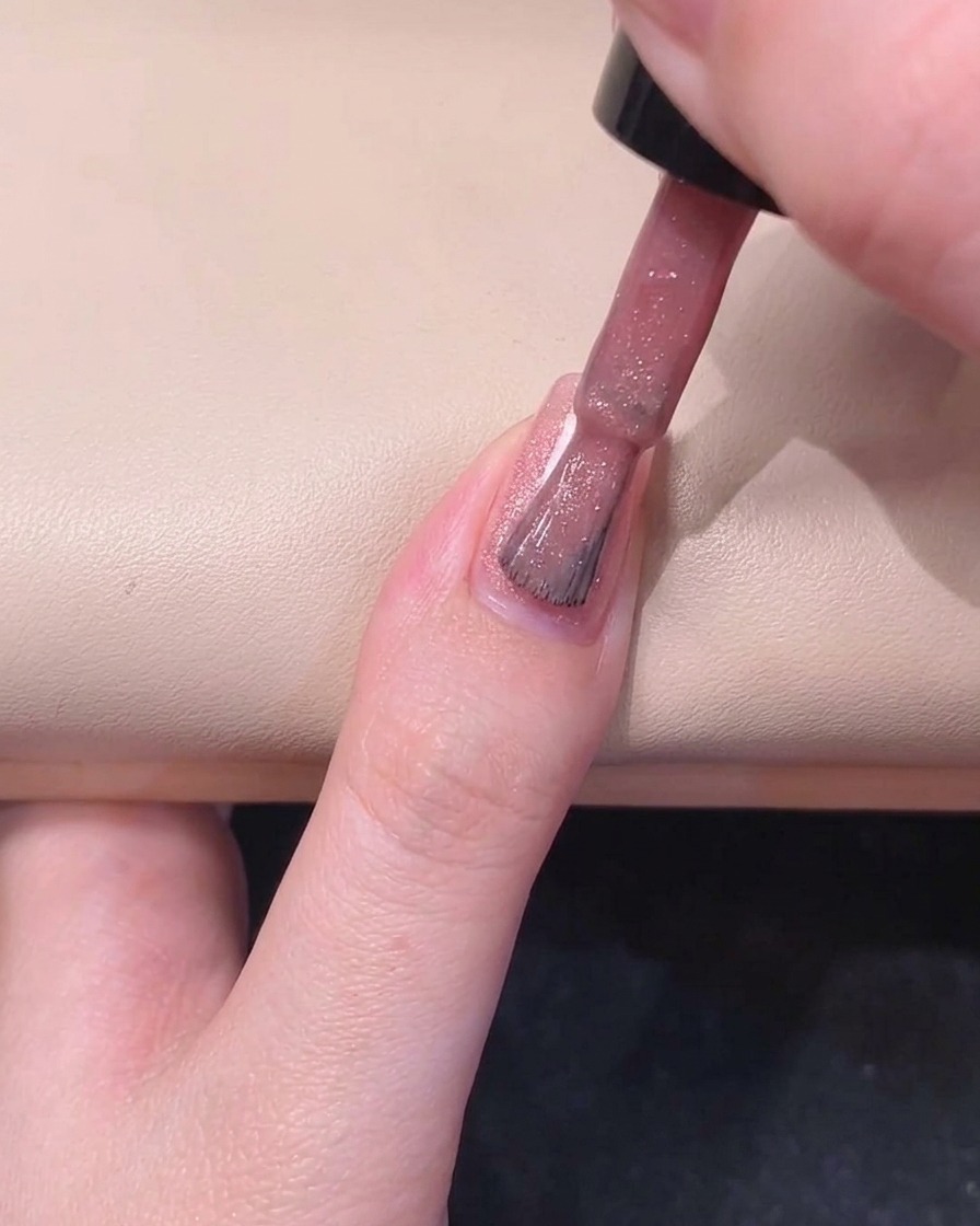 A nail brush painting the right side of a fingernail with shimmering pink gel polish.