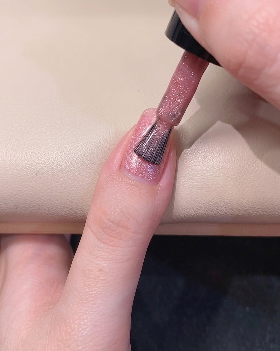 A nail brush spreading sheer pink gel polish along the left side of the fingernail.