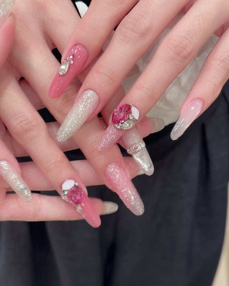 Two perfectly manicured hands resting against a dark fabric background, showcasing finished long pink almond nails with silver glitter gradients and large pink heart rhinestone accents.
