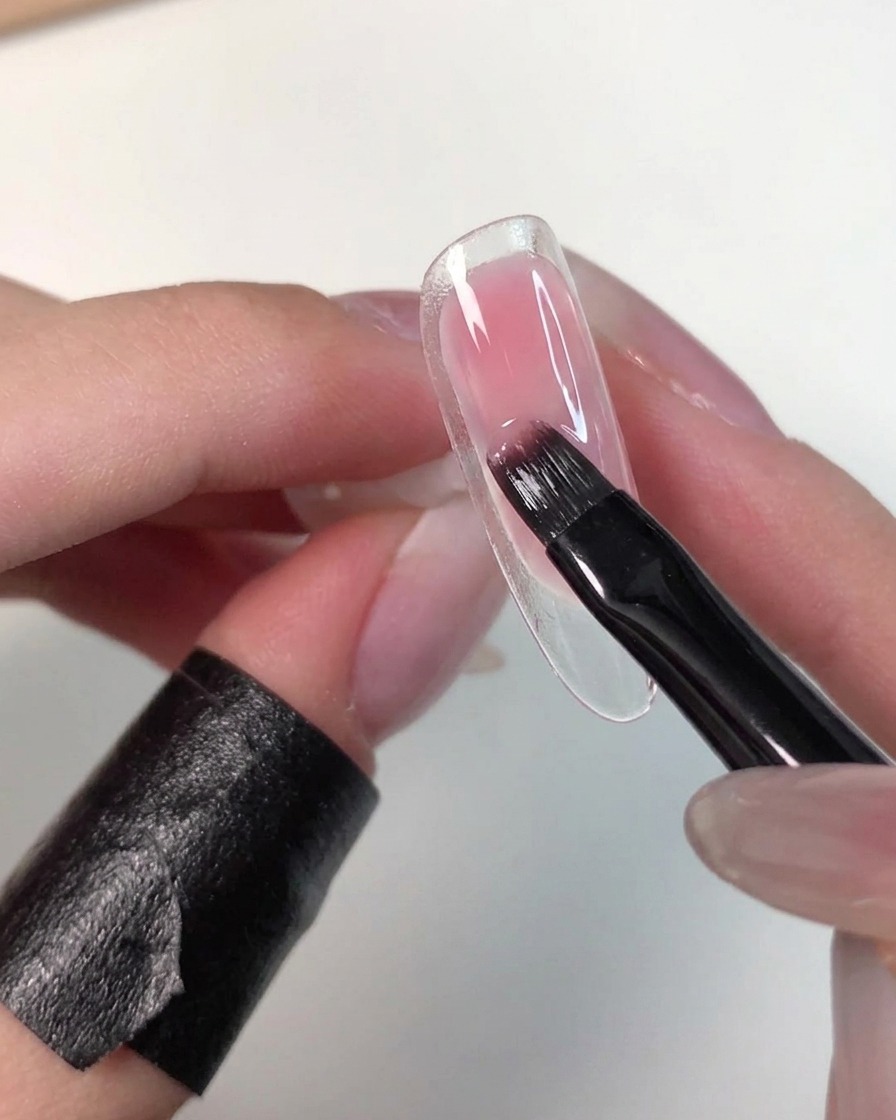 A hand holding a clear nail tip while a flat brush gently blends wet pink gel to soften its edges.