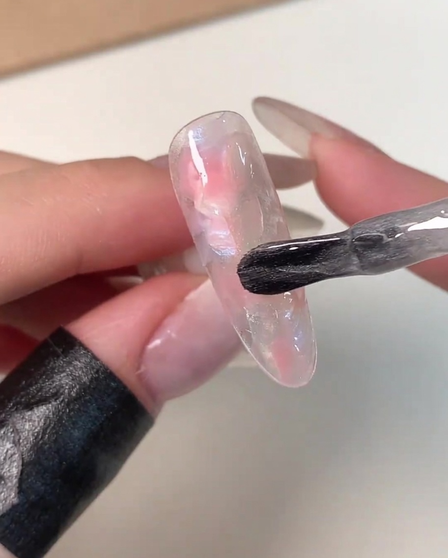 A bottle brush applying a coat of clear, shimmery magnetic gel over a pink and white marbled nail tip.