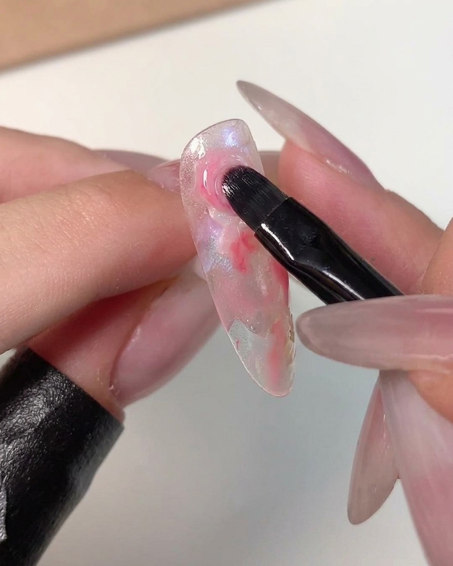 A nail art brush feathering the edges of a dark pink dot into the surrounding lighter colors on a clear nail tip.