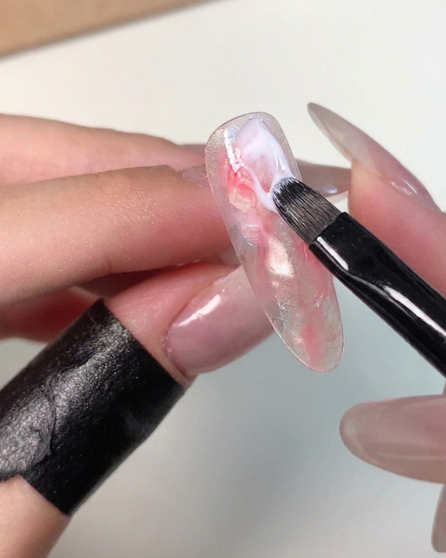 A small gel brush applying a stroke of milky white gel near the cuticle area of a marbled nail tip to refine the pattern.