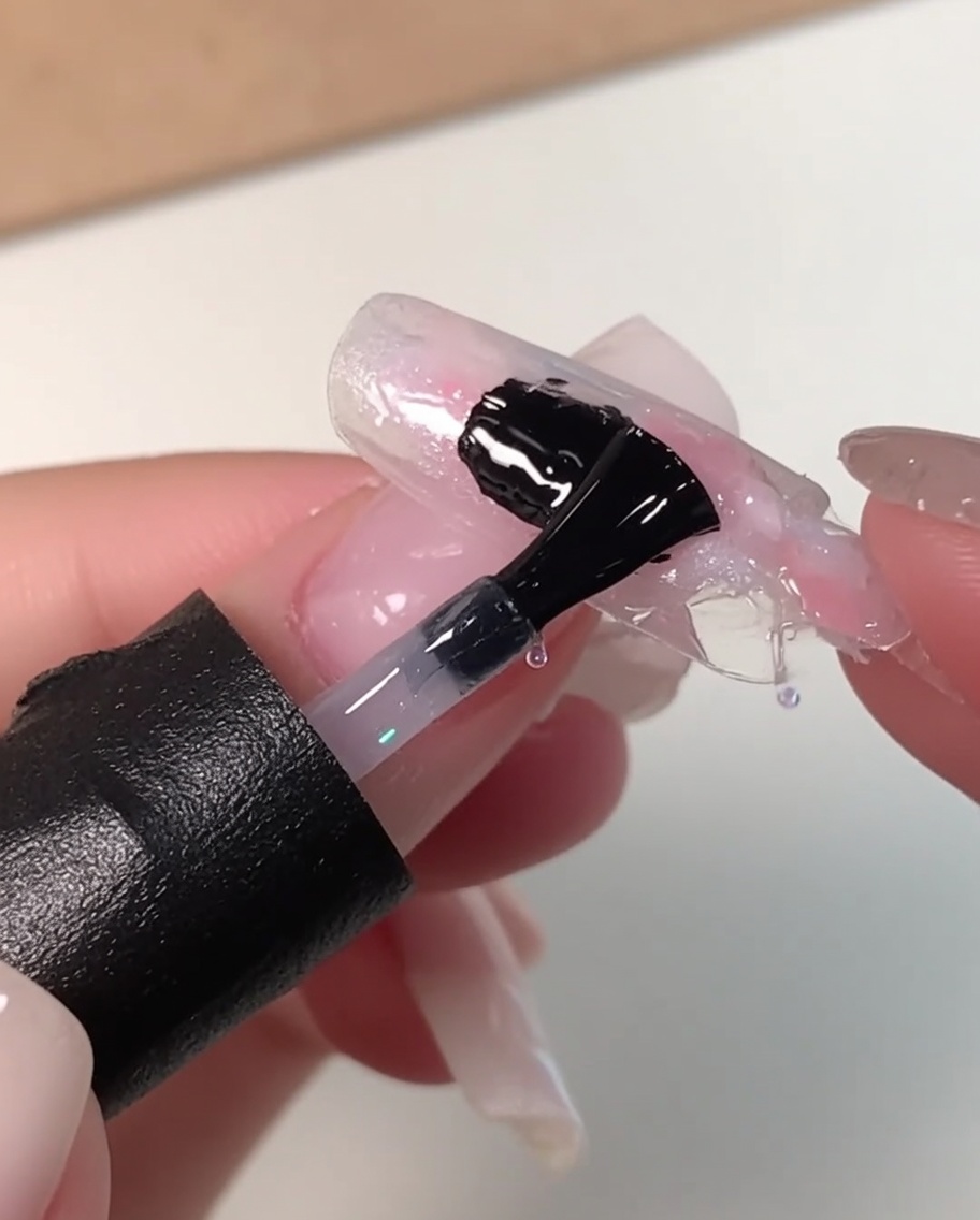 A black nail brush applying a thick layer of clear leveling gel over a marbled pink, white, and blue nail design.
