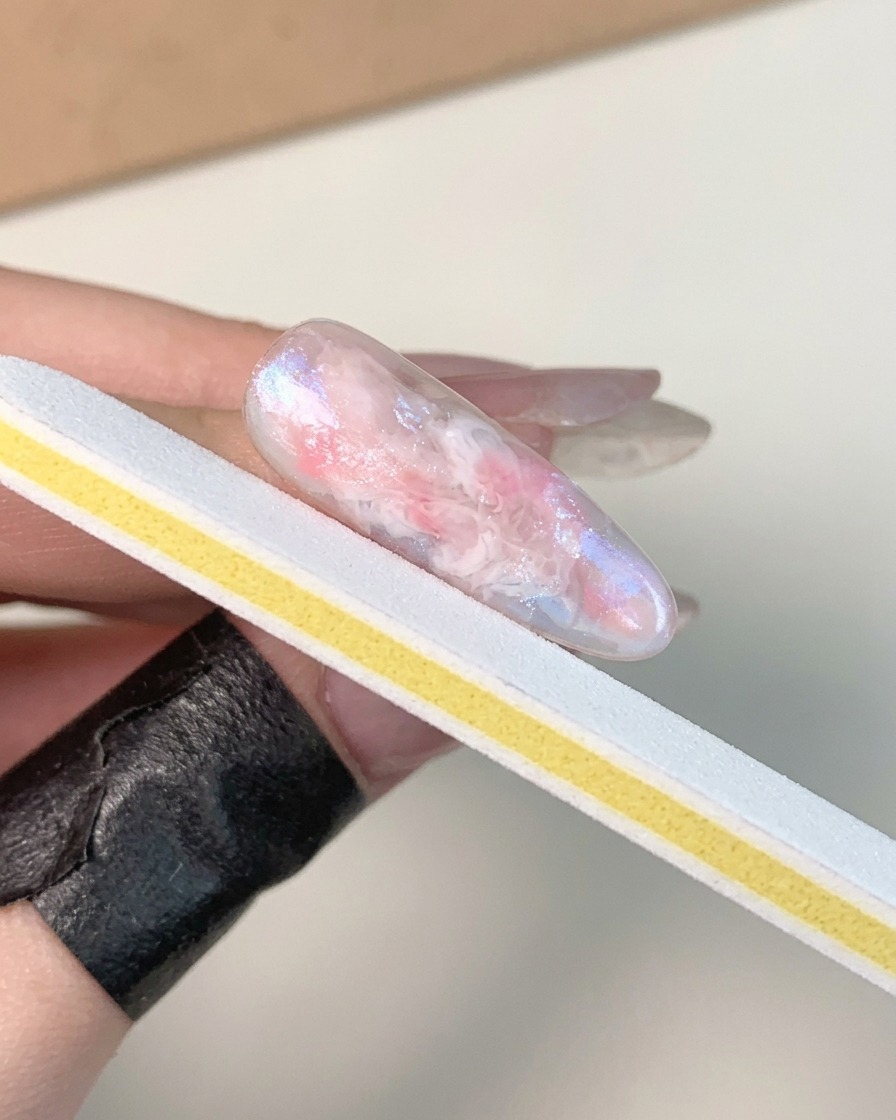 A hand holding a white and yellow nail file strip, buffing the surface of a cured, glossy marbled gel nail.