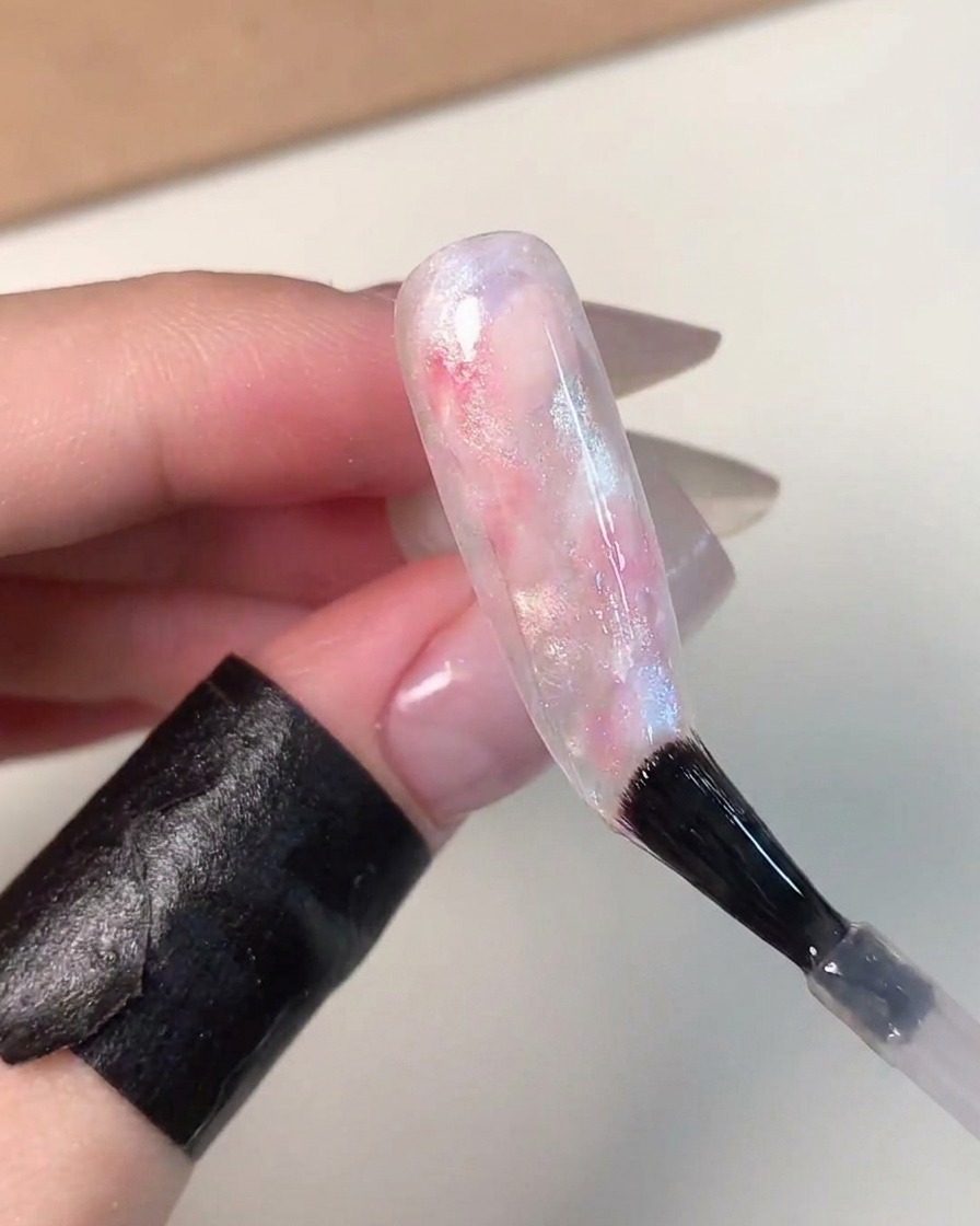A nail brush applying a clear, glossy top coat over a beautifully finished pink, blue, and white marbled glass ball effect nail.