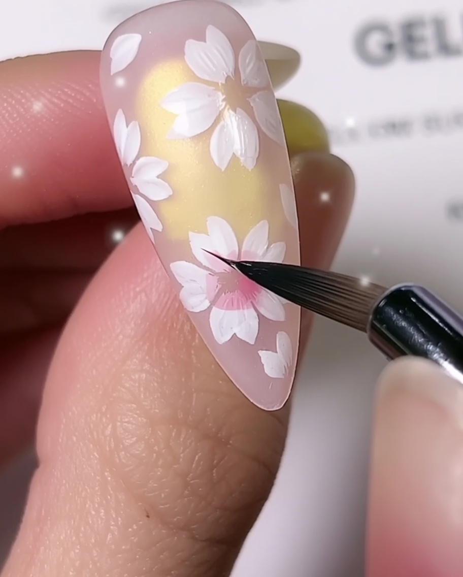 A close-up showing a fine detail nail brush adding pink shading to the center of white cherry blossom petals on an almond-shaped nailcite: 1.