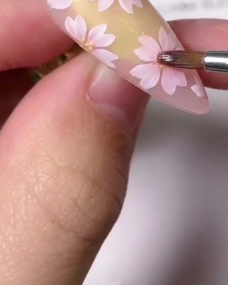 A close-up of a fine detail brush applying small gold center accents to a painted pink cherry blossom on a nail.